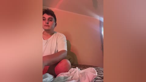 Media: Video of a young, pale-skinned man with short, curly brown hair, wearing a light grey t-shirt and red shorts, sitting on a bed with patterned sheets, under a soft, warm red glow.