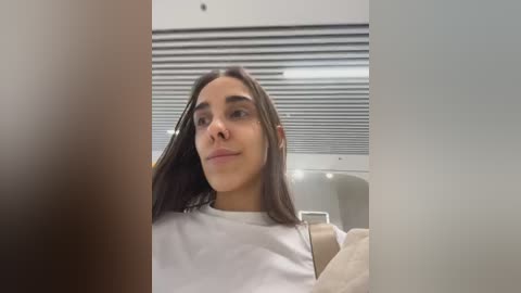 Media: A video of a young woman with long, dark hair, wearing a white T-shirt, seated in a modern indoor setting with white walls and horizontal blinds.