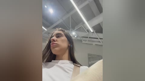 Media: Video of a young woman with long, straight brown hair, wearing a white shirt, standing indoors with exposed ceiling beams and fluorescent lights, looking down with a neutral expression.