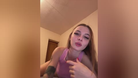 Media: A young woman with long blonde hair, light skin, and a pink striped tank top, puckers her lips and wiggles her fingers in a playful pose, set against a beige-walled room with a wooden door.