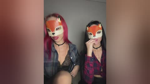 Video of two women with long, straight hair; one with pink hair, one with black. Both wear orange fox masks, black chokers, and plaid shirts. Background is neutral, with a blurred person in the foreground.