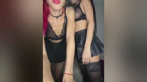 Video of two women in black lingerie and thigh-high stockings, one with vibrant pink hair, standing close together, holding hands, in a dimly lit room.