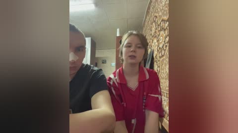 Video of a woman with short brown hair in a black shirt and a young girl with blonde hair in a red polo shirt, standing in a brick-walled hallway with fluorescent lights.