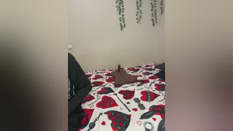 Media: Video of a small, beige dog lying on a bed with a red and black heart-patterned duvet, surrounded by a white room with green ivy hanging from the ceiling.