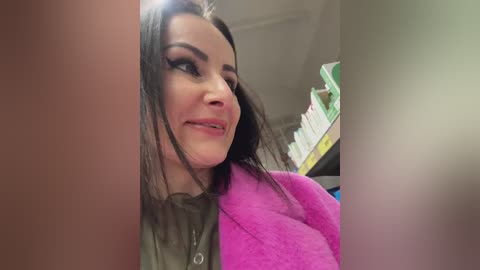 Media: A video of a smiling woman with long dark hair, wearing a pink fleece jacket and a green shirt, standing in a library with bookshelves in the background.