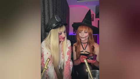Media: Video of two women in Halloween costumes: one with blonde hair and a black top, the other with red hair and a black dress. They are holding a gold-colored object and appear to be in a dimly lit room.
