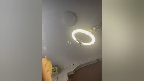 Media: A video of a dimly lit bathroom with a large, glowing circular mirror light above a white sink. The room has a beige wall and a hint of a yellow towel on the left.