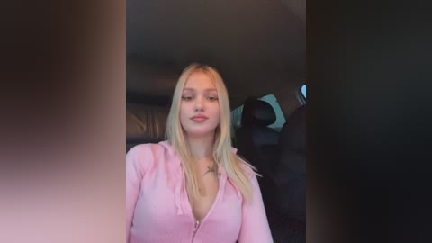 Media: Video of a young Caucasian woman with long blonde hair, wearing a light pink hoodie, sitting in a car. The background features dark interior details and a blue object.