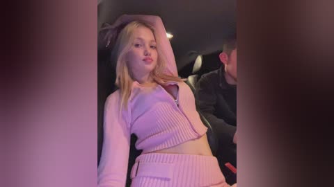 Media: A video of a young blonde woman with fair skin, wearing a pink ribbed crop top and skirt, sitting in a dimly-lit car, looking relaxed.