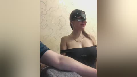 Video of a light-skinned woman with long brown hair in a black mask, black strapless top, and blue jeans, sitting on a textured gray couch in a dimly lit room with patterned wallpaper.