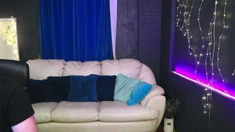 Video of a cozy living room with a cream leather sofa, dark blue and teal throw pillows, and a wall adorned with fairy lights and blue curtains.