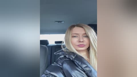 Media: Video of a young, light-skinned blonde woman with straight hair, wearing a black puffer jacket, seated in a car with a gray interior, looking at the camera.