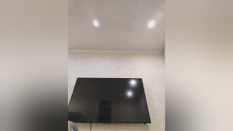 Video of a minimalist, modern room with a large, sleek black flat-screen TV mounted on a beige wall. The ceiling has recessed lighting, casting soft, diffused light. The room's decor is sparse, emphasizing clean lines and neutral tones.
