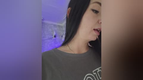 A close-up video of a young woman with long black hair, wearing a gray t-shirt, and a man's erect penis in the background, set against a purple-lit, brick wall.