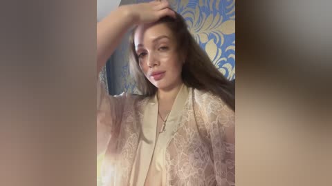 Video of a fair-skinned woman with long, straight brown hair, wearing a sheer, light-colored lace robe, against a blue and gold floral wallpaper background.