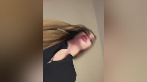 Media: Video of a woman with light skin, long blonde hair, and red lipstick, wearing a black top. She has a slightly blurred, dynamic pose with her head tilted back, against a beige wall.