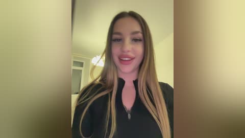 Media: Video of a smiling young woman with long, straight, light brown hair, wearing a black zip-up hoodie. Background features a simple, beige-walled room with a ceiling light fixture.