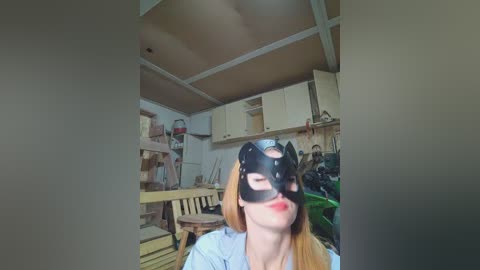 Video of a young woman with fair skin, long blonde hair, and red lipstick, wearing a black cat mask, sitting in a cluttered workshop with wooden furniture and tools.