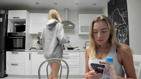 Media: Video of a blonde woman in a grey hoodie and tank top, sitting at a kitchen table, looking at a phone. Another woman in a hoodie cooks in the background.