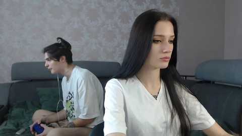 Media: Video of a young woman with long black hair and pale skin, wearing a white shirt, sitting on a dark sofa, frowning. Behind her, a young man with dark hair and glasses, also in a white shirt, sits gaming, headphones on.
