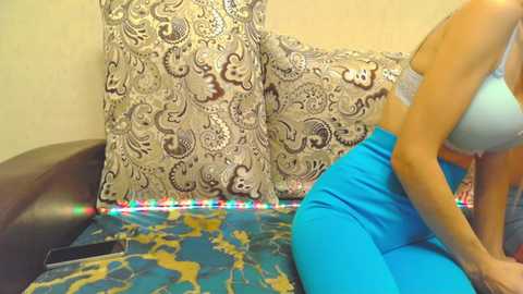 Media: Video of a woman in a light blue sports bra and bright blue leggings, sitting on a blue and gold patterned couch with a black cushion. She is adjusting her bra, and a colorful string of lights is visible on the floor.