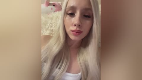 A video of a young woman with long, straight platinum blonde hair, fair skin, and light makeup, wearing a white top, against a beige background. A plush pink bunny is visible in the background.