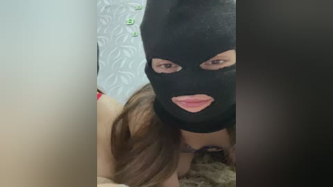 Video of a woman with long brown hair, wearing a black ski mask, lying on a bed with white, floral-patterned sheets.