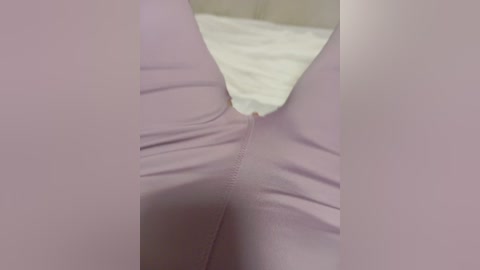 Video of a person's legs in close-up, showing smooth, pale pink leggings with a zipper detail at the crotch. Background includes a white bedsheet and beige pillow.