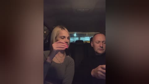 Media: Video of a dimly lit car interior featuring a blonde woman with long hair, wearing a grey sweater, and a bald man in a black shirt. They are seated in the backseat, looking at a smartphone.