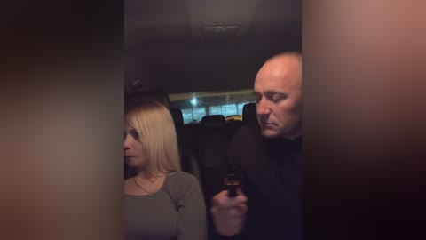 Media: Video of a dimly lit car interior showing a blonde woman in a gray top and a bald man in a black shirt holding a gun, focused on the woman.