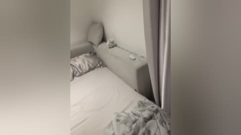 Media: A video of a minimalist, small bedroom with a single bed against a plain white wall. The bed has white sheets and a light gray pillow. A white curtain partially covers the window on the right side. The room is sparsely decorated, giving it a clean, modern feel.