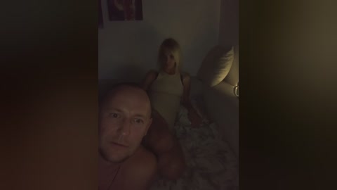 Media: Video of a dimly-lit bedroom featuring a bald man with a beard and a blonde woman with a large chest, both partially clothed, lying on a bed with patterned sheets.