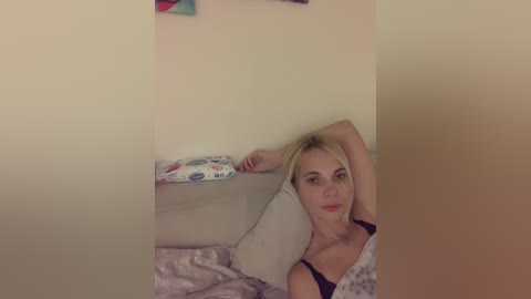 Media: Video of a blonde Caucasian woman with medium build and average breast size, wearing a black top, lying on a beige couch with a floral-patterned blanket, against a cream wall, holding a phone.