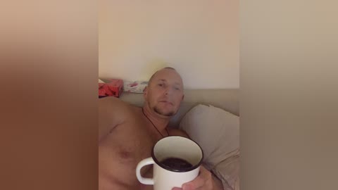 Media: Video of a muscular, bald, bearded man lying shirtless on a beige couch, holding a white mug with black handle, with a plain white wall background.