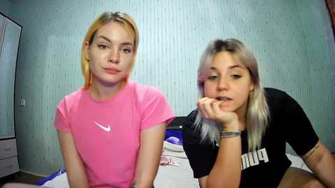 Media: Video of two young women in casual clothing, one with pink Nike shirt and blonde hair, the other with black shirt and long blonde hair, in a bedroom with pastel-colored walls.