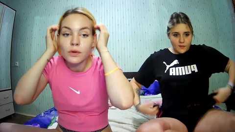 Media: Video of two women in casual clothes: one with blonde hair, wearing a pink top, and the other with dark hair, wearing a black Puma shirt. Both sit on a bed with a blue blanket in a simple room with light blue wallpaper.