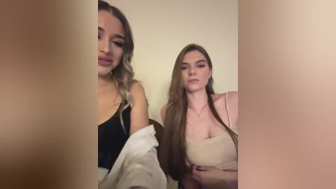 Media: Video of two women: a light-skinned woman with long brown hair in a black top, and a light-skinned woman with long blonde hair in a beige top, both posing against a plain beige background.