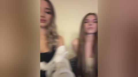 Media: Blurry video of two young women with long blonde hair, one wearing a black top, the other a white garment, both facing a beige wall.