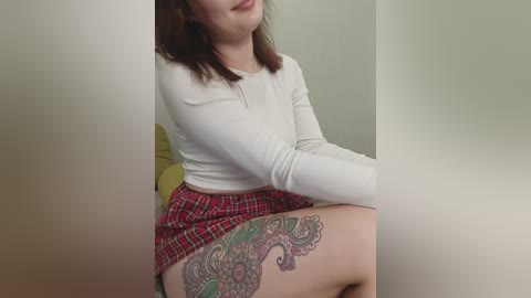 Media: A video of a young woman with light skin, shoulder-length brown hair, and a white long-sleeved top. She sits on a yellow cushioned surface, wearing a red plaid mini-skirt, showcasing a vibrant, detailed floral tattoo on her left thigh.
