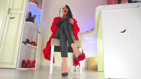 Video of a woman with red lipstick, black hair, red coat, black pants, and black high heels, sitting on a chair in a modern, brightly lit room with white shelves filled with shoes.