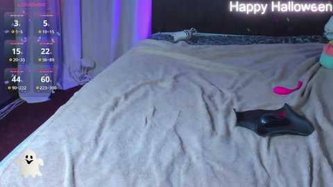 Media: Video of a bedroom with a beige sheet on the bed, a black vibrator, and a pink dildo on the right side. A \"Happy Halloween\" banner hangs on the wall in the background.