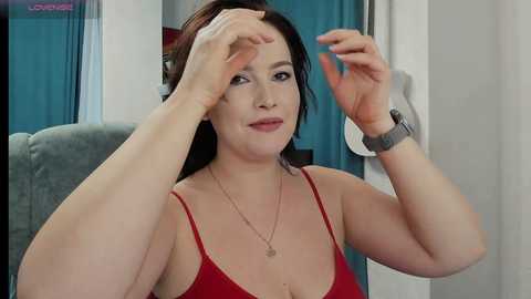 Video of a plus-sized woman with fair skin, dark hair, and red lipstick, adjusting her hair in a mirror. She wears a red spaghetti-strap top and a smartwatch.