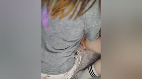 Media: Video of a young woman with long brown hair, wearing a grey sweatshirt, white panties, and striped socks, being embraced from behind by another person, both in a dimly lit room.