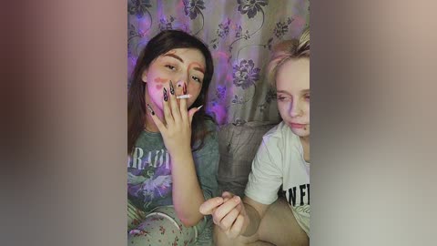 Media: Video of two young women, one smoking, wearing casual clothes against floral-patterned curtains, with a blurred background.