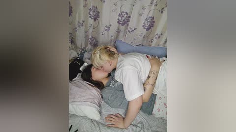 Media: A video shows a blonde woman in a white shirt kissing a brunette woman lying on a bed with floral curtains in the background.