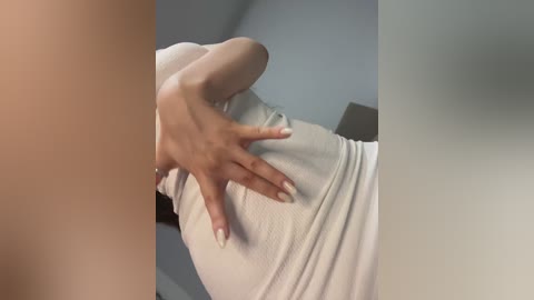 Video of a woman's white, ribbed tank top stretched over her midsection, emphasizing her slender waist and small breasts. Her hand rests on her stomach, fingers splayed. Background features a blurred, grey wall and a piece of furniture.