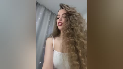 Video of a light-skinned woman with long, curly, blonde hair, wearing a white tank top, posing in a room with a grey curtain and a white ceiling.