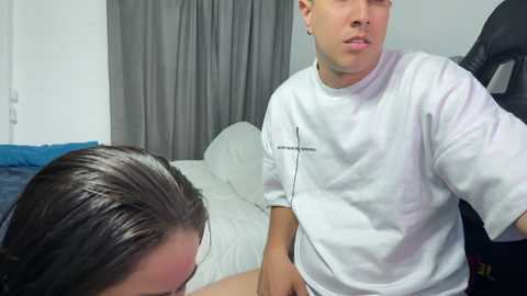 Video of a young man in a white shirt, kneeling beside a woman with wet hair, on a bed with gray curtains in the background.
