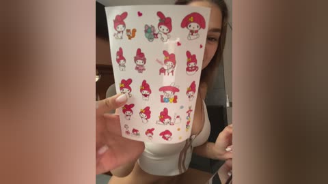 Media: A video of a young woman with light skin, long brown hair, and medium build, holding a white Hello Kitty phone cover with multiple Kitty designs. She wears a white tank top, revealing her midriff. Background shows blurred indoor setting.