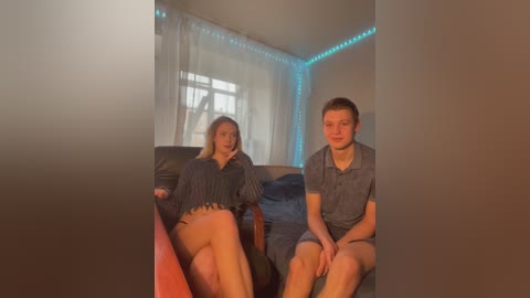 Media: Video of a young Caucasian couple, a pregnant woman in a dark top, and a man in a gray shirt, sitting on a couch in a dimly lit room with blue LED strip lights and a window with sheer curtains.